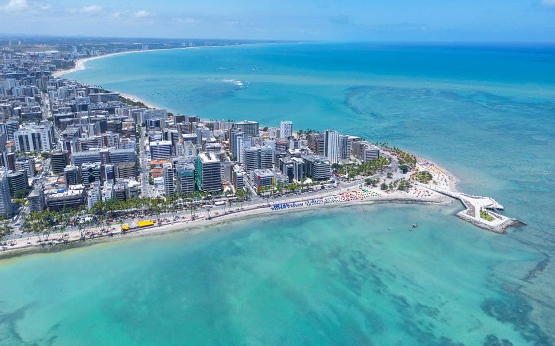 Vista aérea da orla de Maceió com prédios à beira-mar e águas azul-turquesa